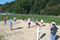 Grupa uczniów wraz z nauczycielami na boisku do piłki siatkowej na piasku. W tle zielona trawa, wysokie drzewa rezerwatu Lisia Góra, po lewej stronie fragment placu zabaw, po prawej ławka
